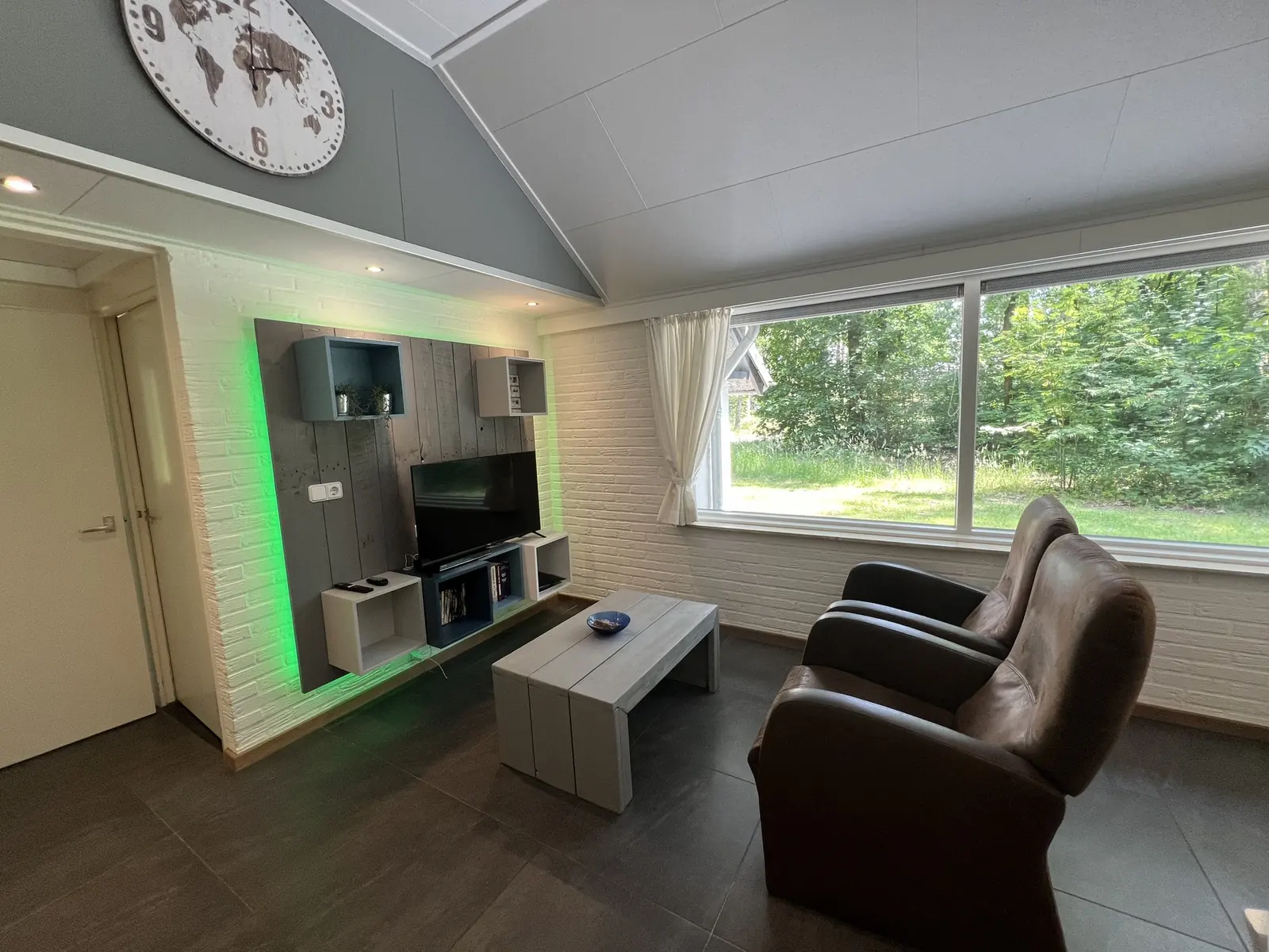 Seating area with two brown armchairs, a TV wall and a large window looking out on the woods