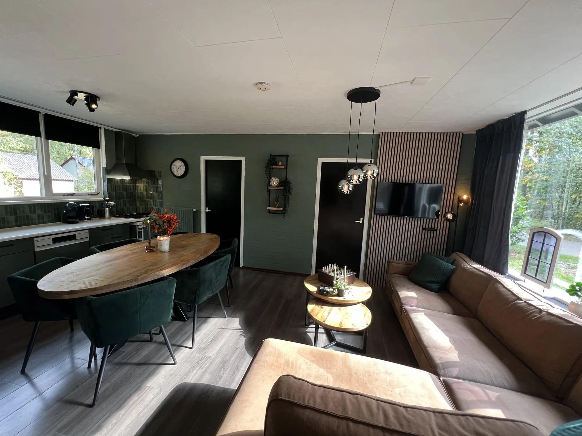 The living room: oval wooden table, green chairs, cognac corner sofa and the kitchen in one space