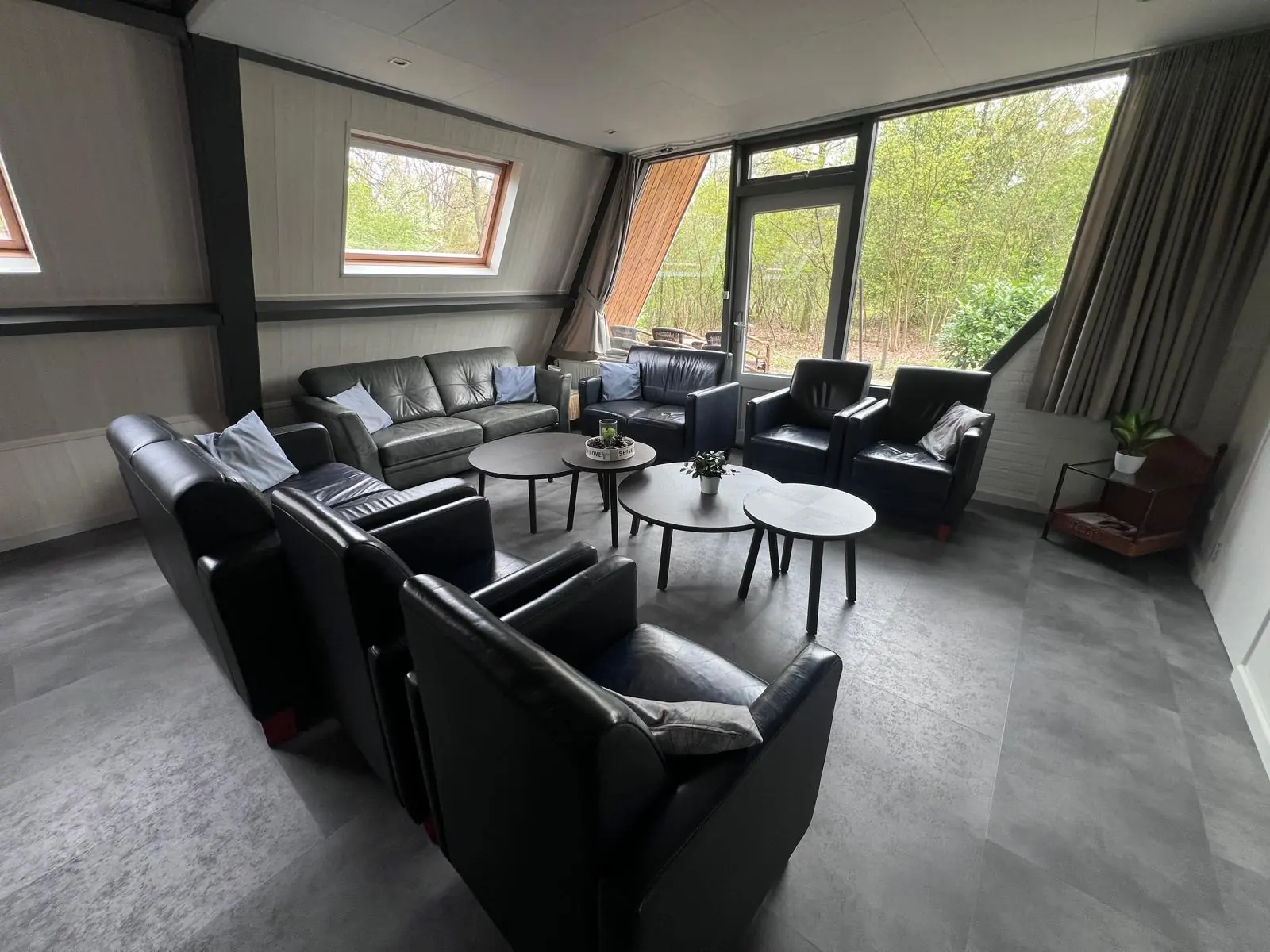 Living room with leather sofas and armchairs around three round tables, windows onto the woods